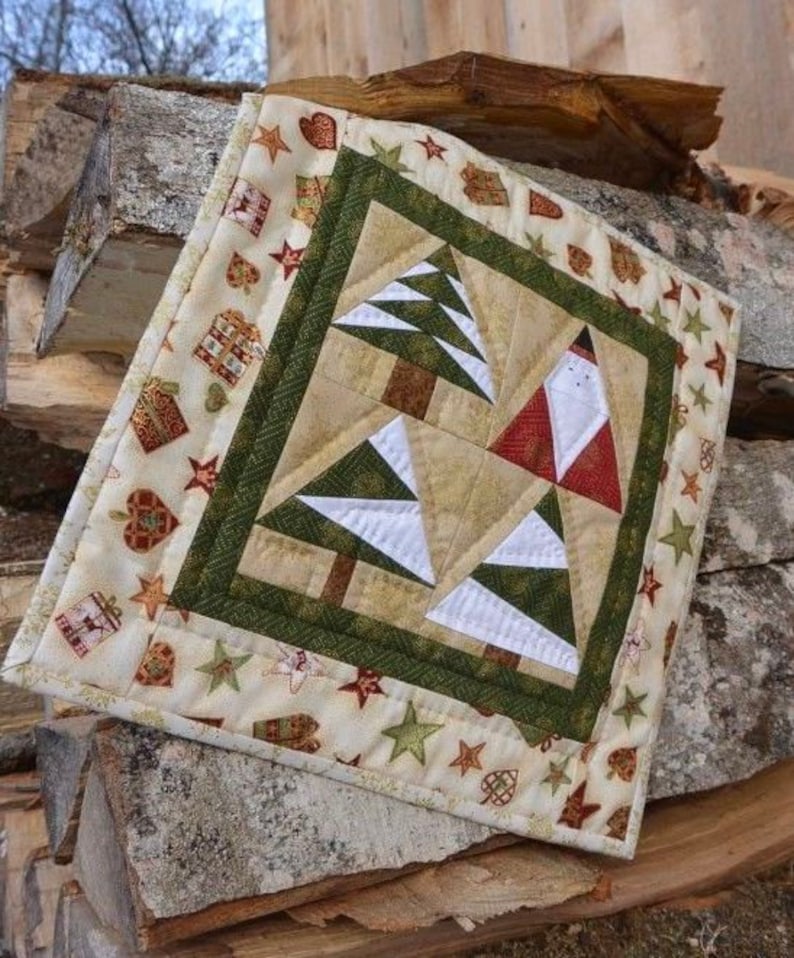 May include: A square Christmas-themed quilted wall hanging. The centre features a patchwork design of Christmas trees and Santa Claus. The border is cream-coloured with repeating patterns of stars, gifts, and hearts. The quilt is displayed on a stack of firewood.