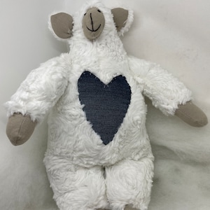 Pode incluir: Um brinquedo de pelúcia de ovelha branca com um patch em forma de coração azul escuro no peito. A ovelha tem orelhas, braços e pés cinzentos. O brinquedo é feito de material macio e fofo e tem um rosto sorridente.