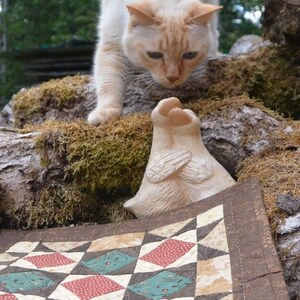 May include: A cream cat with orange markings gazes down at a small, off-white sculpture with wing-like details. A colourful quilt with red, teal, and brown diamond patterns is in the foreground. The scene is set outdoors with moss and tree bark.