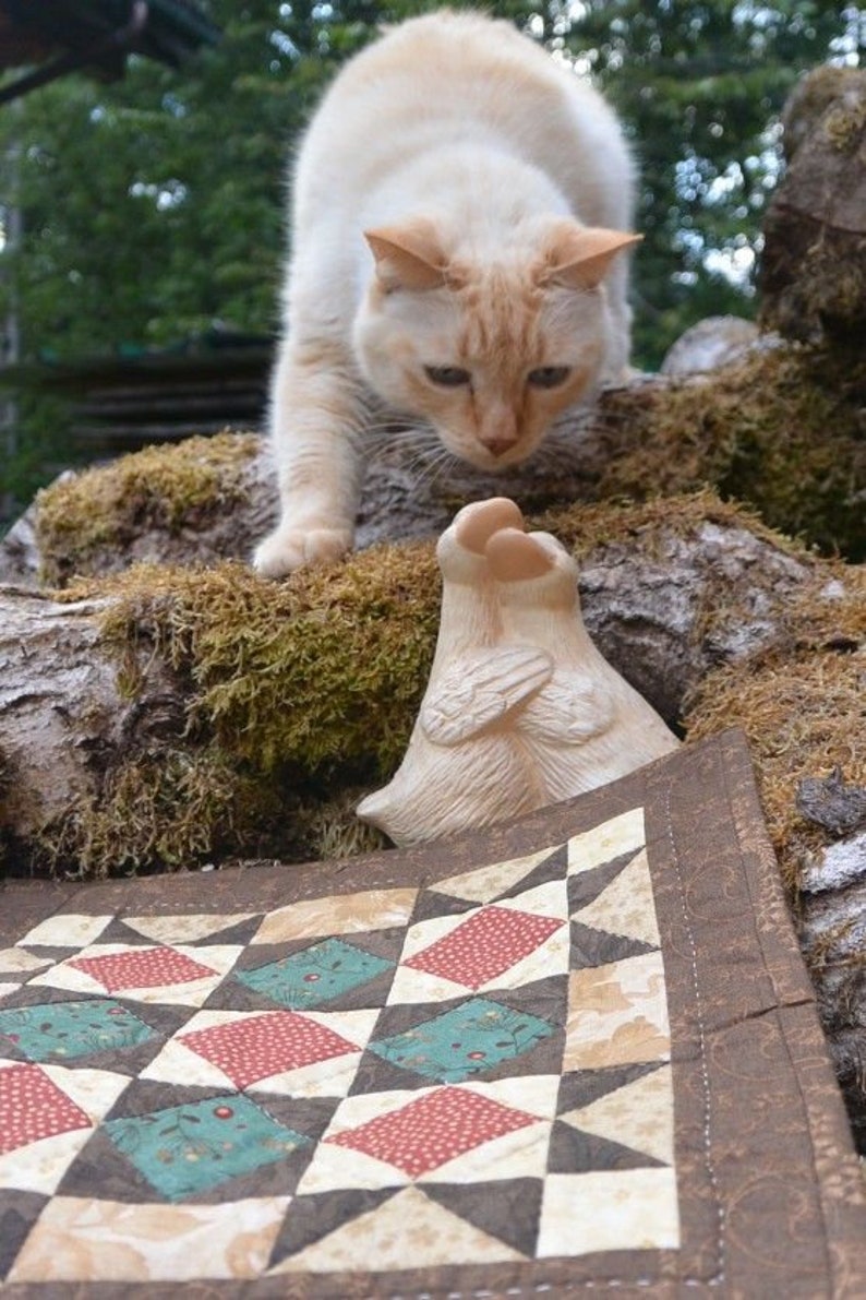 May include: A cream cat with orange markings gazes down at a small, off-white sculpture with wing-like details. A colourful quilt with red, teal, and brown diamond patterns is in the foreground. The scene is set outdoors with moss and tree bark.
