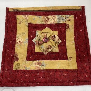 Pode incluir: Bolsa acolchoada quadrada com um design floral em tons de amarelo, vermelho e castanho. O centro apresenta uma flor de tecido com um botão. A bolsa tem fecho de correr e mede aproximadamente 25 cm quadrados.