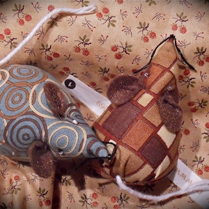 Pode incluir: Dois ratos de tecido feitos à mão. Um rato é azul com um padrão de redemoinho, o outro é castanho com um padrão de losangos. Ambos têm orelhas castanhas e caudas de fio branco. Os ratos estão sobre um tecido com padrão floral.