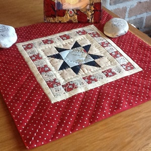 Op de afbeelding: Een handgemaakte, gewatteerde tafelloper met een rood stippenpatroon en een centraal sterontwerp. De ster is samengesteld uit blauwe, cr&egrave;mekleurige en zwarte stof. Twee stenen met tekst staan aan weerszijden van de loper.