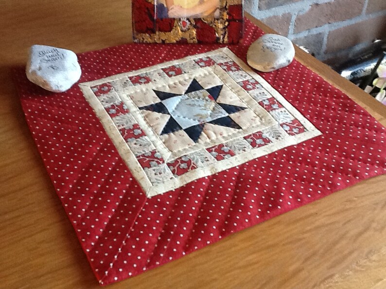 Op de afbeelding: Een handgemaakte, gewatteerde tafelloper met een rood stippenpatroon en een centraal sterontwerp. De ster is samengesteld uit blauwe, cr&egrave;mekleurige en zwarte stof. Twee stenen met tekst staan aan weerszijden van de loper.