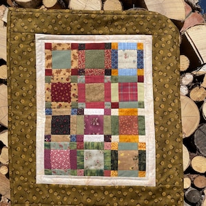 Peut inclure: Une courtepointe patchwork faite à la main avec une variété de carrés de tissu colorés. La courtepointe présente un motif central de carrés dans des tons de rouge, vert, bleu et marron, encadré d'une bordure crème et d'un bord vert olive à motifs.