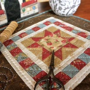 Op de afbeelding: Een handgemaakte, gewatteerde onderzetter met een sterpatroon in rode, gouden en blauwe stoffen. De onderzetter is ruitvormig, met een bloemenmidden en een decoratieve rand. Een antieke schaar ligt op de onderzetter.