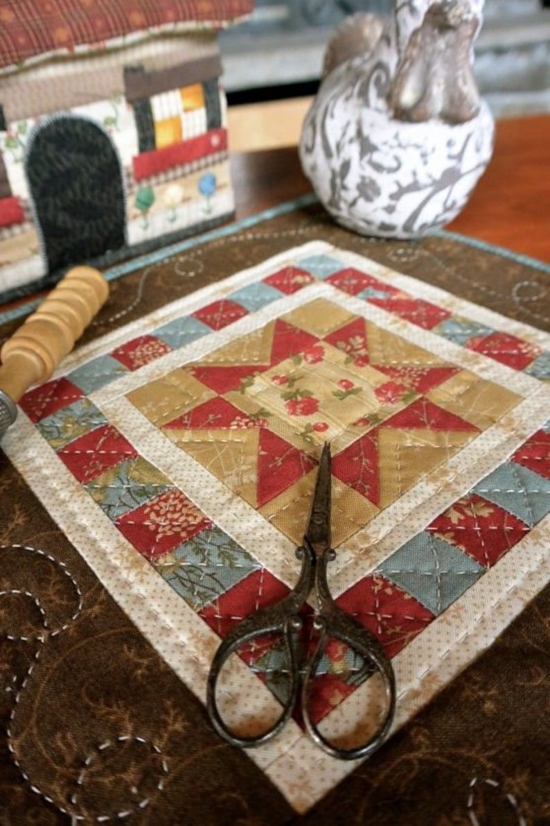 Op de afbeelding: Een handgemaakte, gewatteerde onderzetter met een sterpatroon in rode, gouden en blauwe stoffen. De onderzetter is ruitvormig, met een bloemenmidden en een decoratieve rand. Een antieke schaar ligt op de onderzetter.