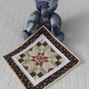 Pu&ograve; includere: Un orsacchiotto patchwork fatto a mano nei toni del blu, con un tappetino quadrato trapuntato abbinato. Il tappetino presenta un motivo a stella con tessuto rosso, verde e marrone e un bordo floreale. L'orso &egrave; su una superficie bianca.