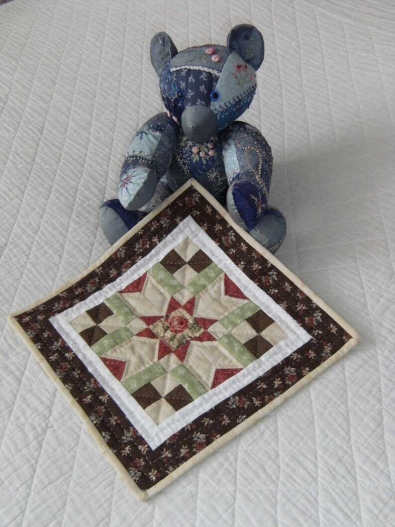 Pu&ograve; includere: Un orsacchiotto patchwork fatto a mano nei toni del blu, con un tappetino quadrato trapuntato abbinato. Il tappetino presenta un motivo a stella con tessuto rosso, verde e marrone e un bordo floreale. L'orso &egrave; su una superficie bianca.