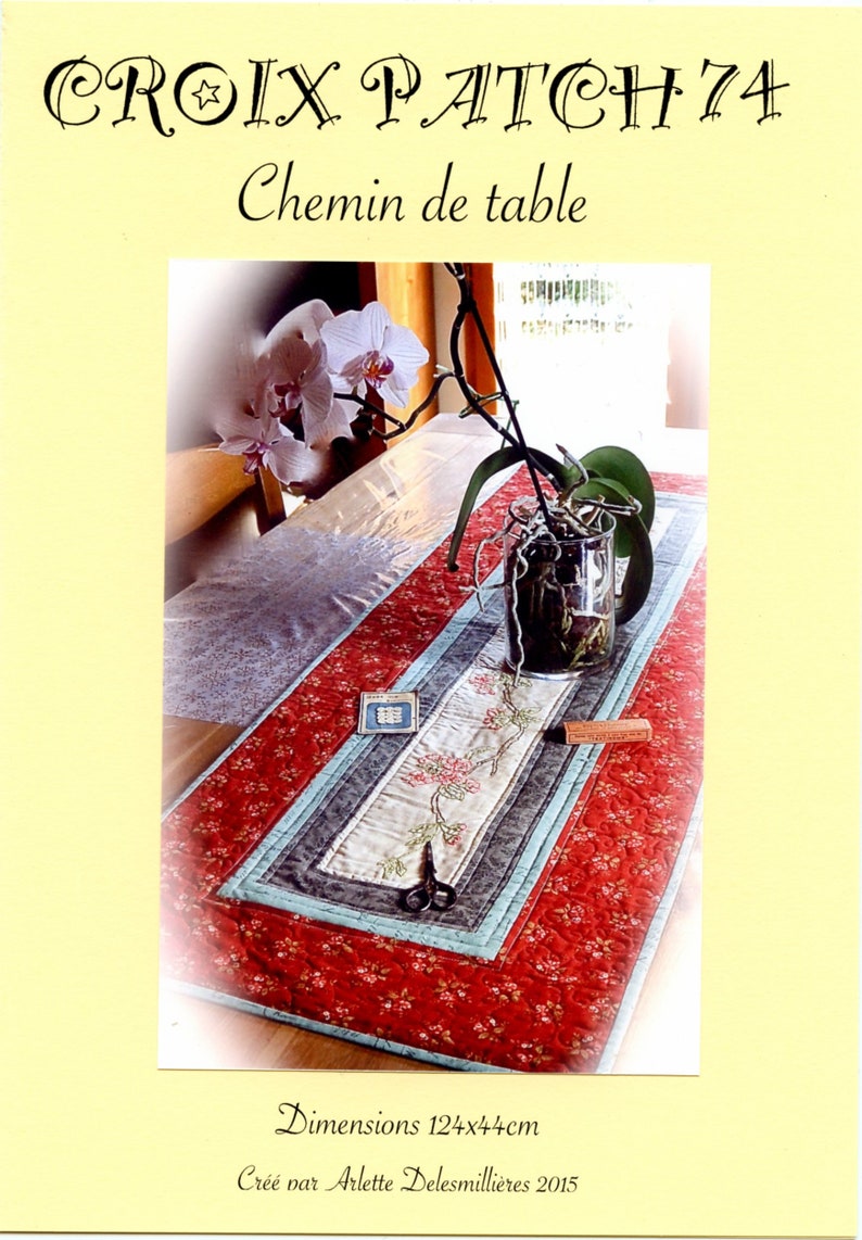 Peut inclure: Chemin de table rouge &agrave; motifs floraux, avec un motif floral brod&eacute; au centre, bord&eacute; de rayures bleues et grises. Le chemin de table mesure 124x44cm. Le texte "CROIX PATCH 74" et "Chemin de table" sont en haut.