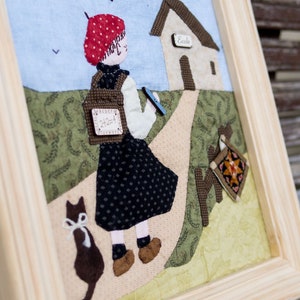 Peut inclure: Une œuvre d'art encadrée en tissu représentant un enfant marchant vers une école. L'enfant porte un chapeau rouge à pois et porte un sac à dos avec un problème de mathématiques. Un chat et le mot "Ecole" sont également visibles.