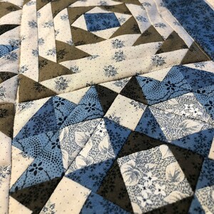 K&ouml;nnte beinhalten: Nahaufnahme eines handgefertigten Quilts mit geometrischem Muster. Der Quilt zeigt Quadrate und Dreiecke in Blau-, Creme- und Graut&ouml;nen. Der Stoff hat Blumen- und Punktmuster mit sichtbaren N&auml;hten im gesamten Design.