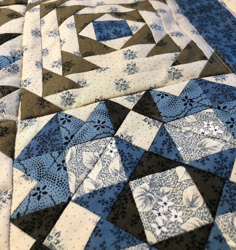 K&ouml;nnte beinhalten: Nahaufnahme eines handgefertigten Quilts mit geometrischem Muster. Der Quilt zeigt Quadrate und Dreiecke in Blau-, Creme- und Graut&ouml;nen. Der Stoff hat Blumen- und Punktmuster mit sichtbaren N&auml;hten im gesamten Design.