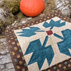 May include: An orange gourd sits on moss beside a quilted placemat. The placemat has a central design of teal leaf shapes on a beige background, framed by a brown border with leaf patterns.
