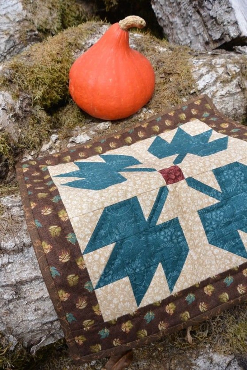 May include: An orange gourd sits on moss beside a quilted placemat. The placemat has a central design of teal leaf shapes on a beige background, framed by a brown border with leaf patterns.