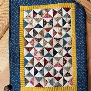 Peut inclure: Une courtepointe faite à la main avec un motif patchwork. La courtepointe présente une section centrale de triangles alternés dans divers motifs et couleurs, dont le rouge, le bleu et le marron. Elle est bordée d'une bande jaune et d'un tissu bleu marine avec un petit motif floral.