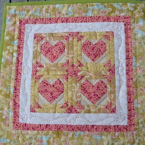 May include: A handmade quilt with a floral pattern in shades of yellow, pink, and green. The center features four heart-shaped designs. The quilt is bordered with a green edge and white decorative stitching.