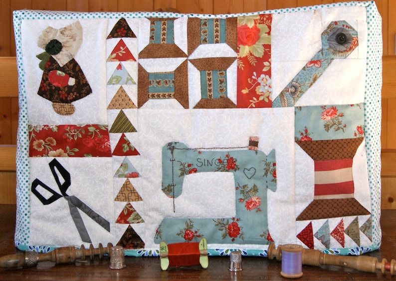 K&ouml;nnte beinhalten: Ein Patchwork-Quilt mit N&auml;h-Motiven. Das Design beinhaltet eine N&auml;hmaschine, Garnrollen, eine Schere und eine Figur mit Haube. Die Farbpalette umfasst Wei&szlig;, T&uuml;rkis, Rot und Braun. Das Wort "SINGER" ist auf der N&auml;hmaschine zu sehen.