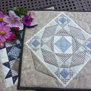 May include: A patchwork quilt square with a geometric pattern in blue, grey, and white. The square has a central diamond shape with smaller squares and triangles. A bouquet of pink and purple flowers is placed nearby.