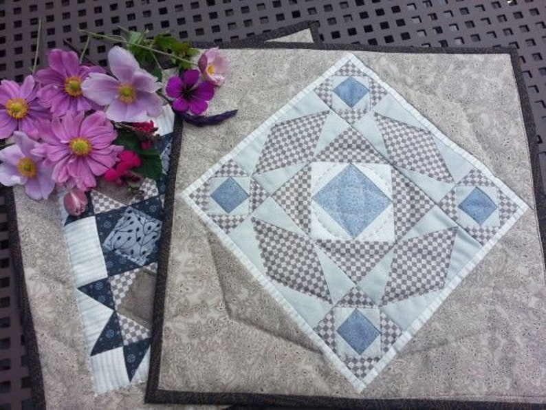 May include: A patchwork quilt square with a geometric pattern in blue, grey, and white. The square has a central diamond shape with smaller squares and triangles. A bouquet of pink and purple flowers is placed nearby.