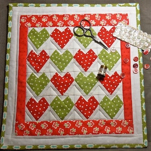 Pode incluir: Um quadrado acolchoado feito à mão com um padrão de corações repetidos em bolinhas vermelhas e verdes sobre um fundo branco. O edredom é contornado com tecido floral vermelho e uma borda estampada em verde e branco. Materiais de costura, incluindo tesoura, botões e dedal, são colocados próximos.