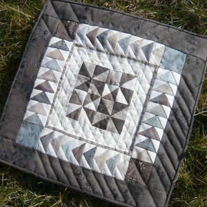 May include: A square, handmade quilt with a geometric design. The quilt features a central starburst pattern in shades of gray and white, surrounded by borders of triangles and a dark gray edge. The quilt is placed on a bed of green grass.
