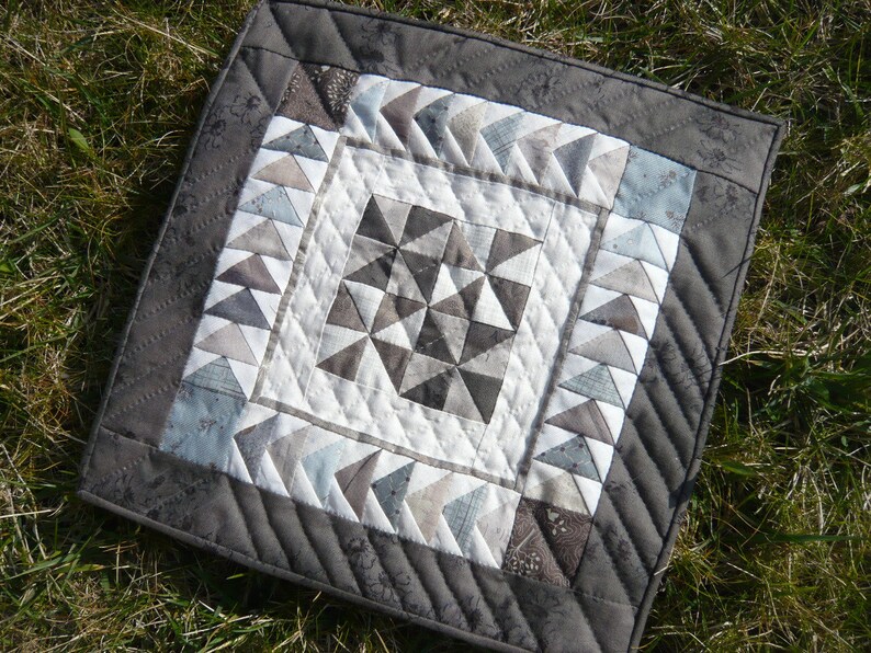 May include: A square, handmade quilt with a geometric design. The quilt features a central starburst pattern in shades of gray and white, surrounded by borders of triangles and a dark gray edge. The quilt is placed on a bed of green grass.