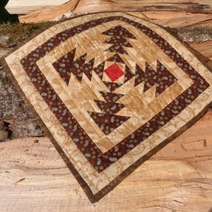 May include: A square quilted placemat with a pineapple design. The placemat features a central red square, surrounded by brown and tan triangles. The border is a paisley pattern in brown and tan.