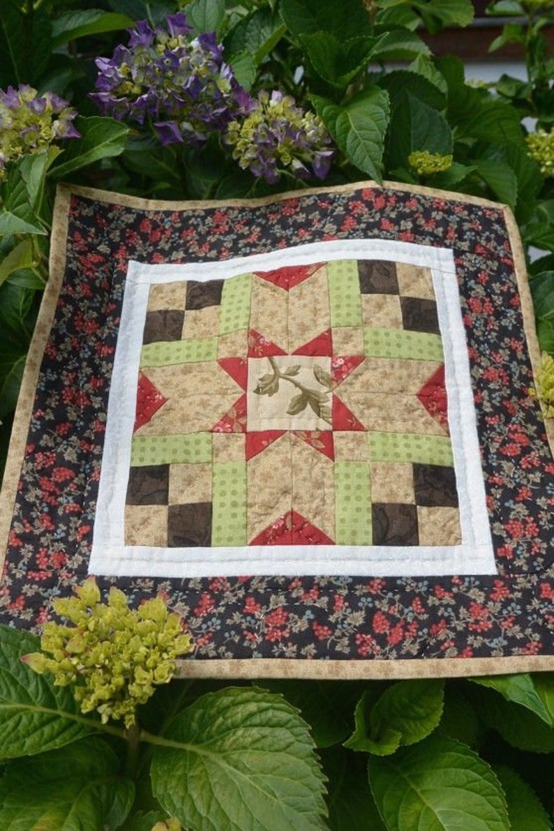 Pu&ograve; includere: Una piccola trapunta quadrata con un motivo a stella centrale. La trapunta presenta un bordo bianco, un bordo esterno a fantasia floreale e un motivo patchwork nei toni del beige, verde, rosso e marrone. Circondata da fogliame e fiori verdi.