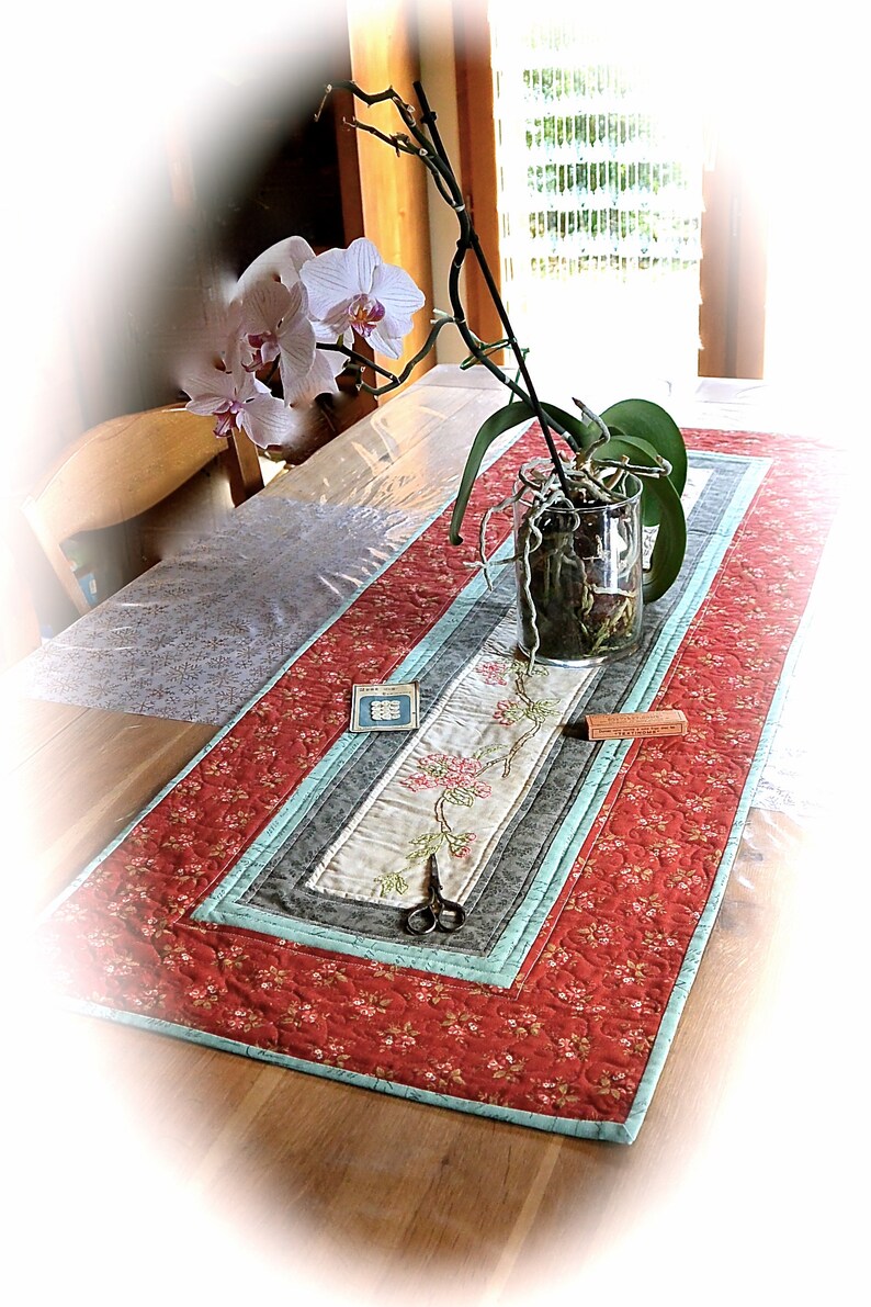 Peut inclure: Chemin de table matelass&eacute; avec un motif floral rouge, gris et turquoise. Le panneau central pr&eacute;sente des fleurs brod&eacute;es. Un vase en verre transparent avec une plante d'orchid&eacute;e est pos&eacute; sur le chemin de table. Des ciseaux et de petits objets sont &eacute;galement pr&eacute;sents.