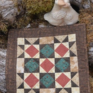 May include: A small, square quilt with a patchwork design featuring red, teal, and cream-colored diamond and star patterns. The quilt is bordered with a brown floral fabric. A ceramic duck figurine is perched above the quilt.