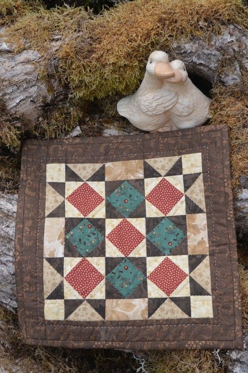 May include: A small, square quilt with a patchwork design featuring red, teal, and cream-coloured diamond and star patterns. The quilt is bordered with a brown floral fabric. A ceramic duck figurine is perched above the quilt.