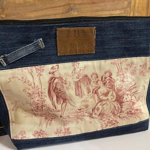 Pode incluir: Bolsa tote de ganga com fecho de correr preto e remendo decorativo. A bolsa apresenta um painel creme com uma cena toile de Jouy vermelha e branca. Um remendo de couro castanho com a palavra "Doine" é costurado na ganga.