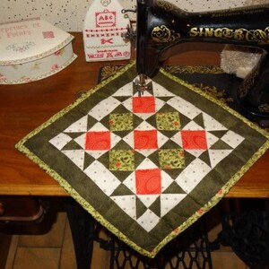May include: A square, quilted potholder with a geometric design in red, green, white, and black. It's placed on a wooden table next to a vintage Singer sewing machine. The potholder is approximately 20 cm square.
