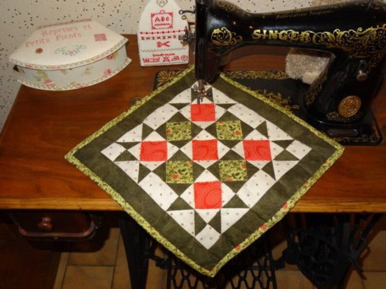May include: A square, quilted potholder with a geometric design in red, green, white, and black. It's placed on a wooden table next to a vintage Singer sewing machine. The potholder is approximately 20 cm square.