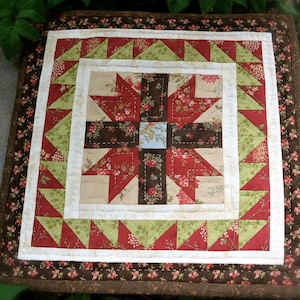 May include: A square quilted table topper with a floral pattern. The centre features a cross design in red, brown, and beige fabrics. The border includes green and red triangles, with a floral patterned edge.