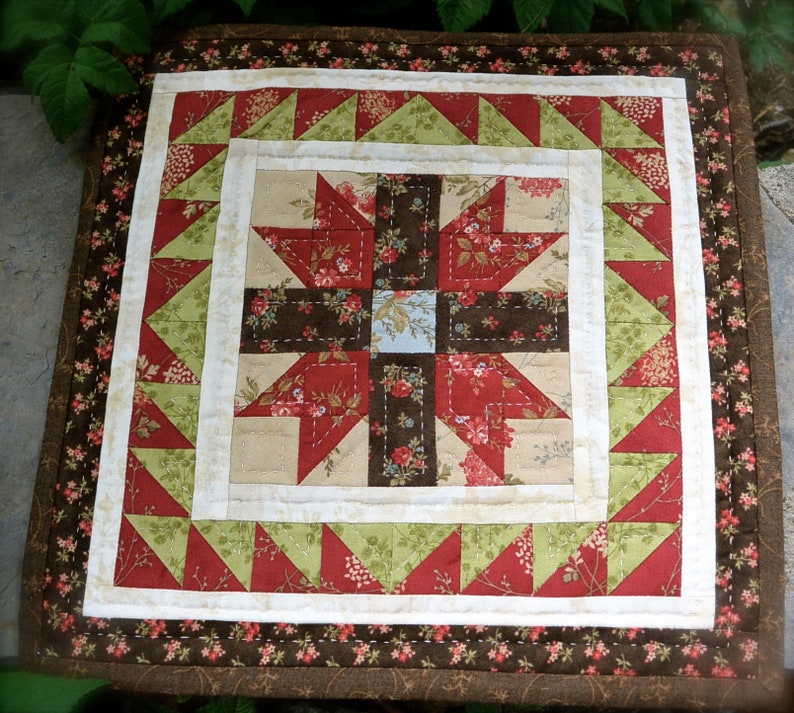 May include: A square quilted table topper with a floral pattern. The centre features a cross design in red, brown, and beige fabrics. The border includes green and red triangles, with a floral patterned edge.