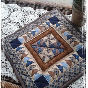 May include: A square quilted placemat with a brown border and a central diamond pattern. The design features triangles in shades of blue, tan, and brown, creating a patchwork effect. The placemat is placed on a wicker surface.