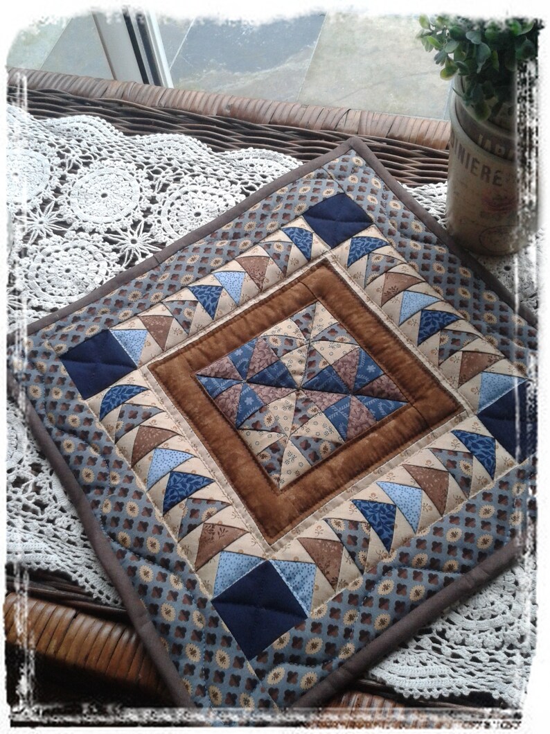 May include: A square quilted placemat with a brown border and a central diamond pattern. The design features triangles in shades of blue, tan, and brown, creating a patchwork effect. The placemat is placed on a wicker surface.