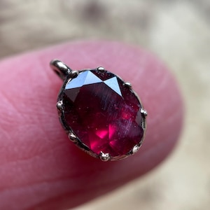 May include: A faceted, oval-shaped, deep red gemstone pendant. The gem is set in a silver-colored metal with a small loop at the top for a chain. The pendant is held between two fingers.