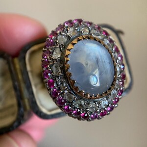 May include: An oval-shaped ring with a large, light blue center stone, surrounded by a halo of small, pink and clear gemstones. The ring has a gold-colored setting and is displayed in a vintage jewelry box.