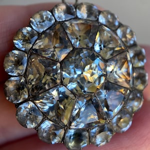 May include: A round, antique pendant with a cluster of clear, faceted gemstones. The gemstones are set in a silver-toned metal frame, creating a sparkling, floral-like design. The pendant has a small loop at the top for hanging.