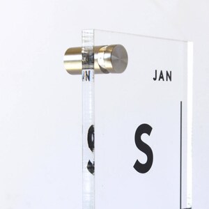 May include: A clear acrylic calendar with a black grid and the word "JAN" printed in black. The calendar is mounted on a wall with a silver metal bracket.