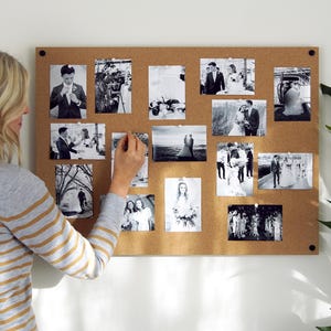 Op de afbeelding: Een kurkboard met zwart-witfoto's van een bruidspaar die erop geprikt zijn. De foto's laten het paar zien terwijl ze zich klaarmaken, foto's maken en dansen op hun bruiloft.