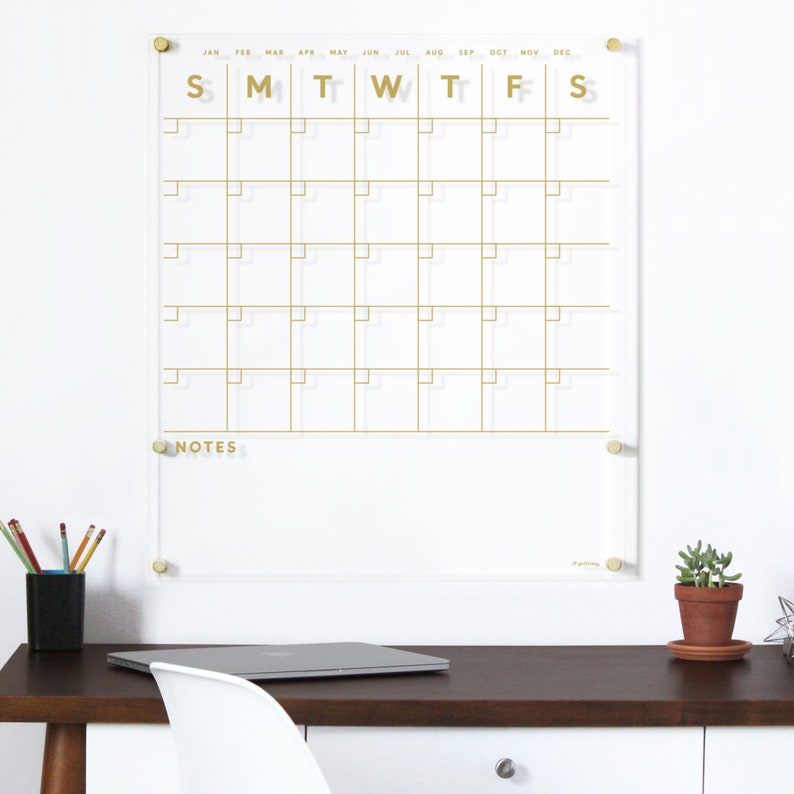 Op de afbeelding: Een transparante, droog uitwisbare acrylkalender met gouden letters en een gouden frame. De kalender heeft een raster voor elke maand van het jaar en een sectie voor notities. De kalender hangt aan een witte muur boven een bureau met een laptop en een potplant.
