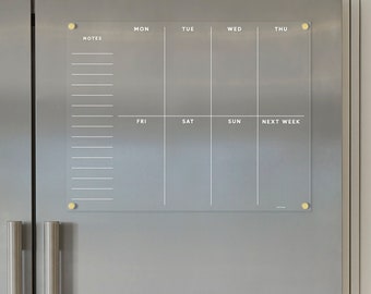 以下が含まれることがあります： 金色のプッシュピンが付いた透明なアクリル製の週間プランナー。プランナーには左側にメモ欄があり、週の各曜日と「来週」の欄があります。