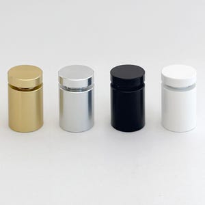 Op de afbeelding: Vier cilindrische displaystandaards in goud, zilver, zwart en wit. Elke standaard heeft een kleine transparante schijf bovenop om items te presenteren.