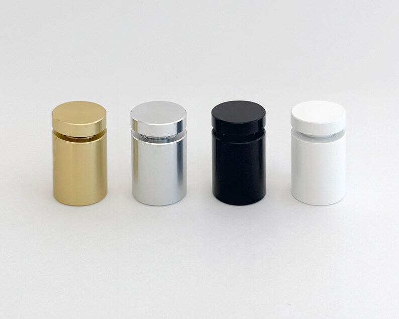 Op de afbeelding: Vier cilindrische displaystandaards in goud, zilver, zwart en wit. Elke standaard heeft een kleine transparante schijf bovenop om items te presenteren.