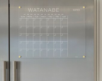 May include: A clear acrylic dry-erase calendar with the name "WATANABE" at the top and the months of the year listed across the top row. The days of the week are listed across the second row. The calendar is mounted on a stainless steel refrigerator.