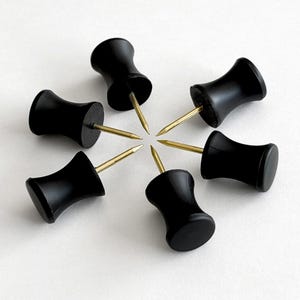 May include: Six black push pins arranged in a circular pattern. Each pin has a rounded, hourglass-shaped head and a gold-colored pin. The pins are set against a white background.